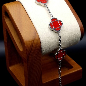 Puede incluir: Una pulsera de plata con tres colgantes rojos en forma de trébol con bordes plateados, unidos por una fina cadena. La pulsera se exhibe en un expositor de joyas de madera con un cojín color crema.