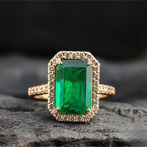 Puede incluir: Un anillo de oro con una gran esmeralda verde de corte rectangular. La esmeralda está rodeada por un halo de piedras preciosas redondas y marrones más pequeñas. La banda también está adornada con pequeñas piedras redondas.