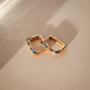 May include: A pair of gold-colored, square hoop earrings. Each earring is adorned with a row of colorful gemstones, including blue, green, red, and yellow. The earrings are displayed on a light brown surface, with soft lighting.