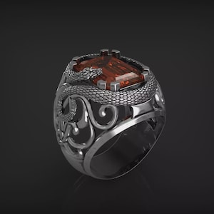 Peut inclure: Une bague en argent avec une pierre précieuse rectangulaire brun-rouge. La bague présente un motif de serpent détaillé enroulé autour de la pierre, avec des volutes ornementales sur le bandeau. La bague est posée sur un fond sombre.