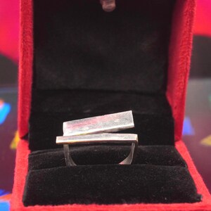 May include: A silver ring featuring two parallel, rectangular metal bars positioned horizontally across the top. The ring is displayed inside a bright red jewelry box with a black velvet interior lining that holds the band securely in place.