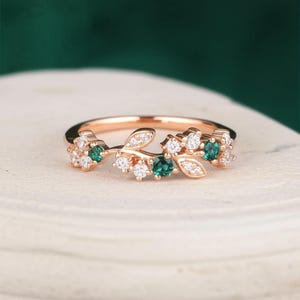 May include: A rose gold ring with a leaf design. The ring features small, round, emerald green gemstones and clear, round gemstones. The ring is set on a white surface with a dark green background.