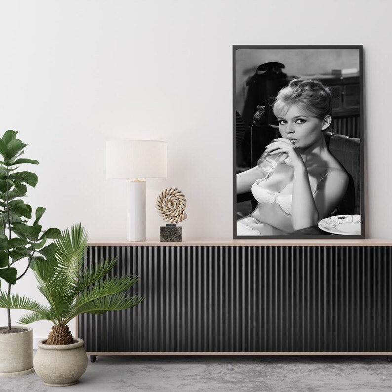 Puede incluir: Impresi&oacute;n en blanco y negro enmarcada de una mujer con sujetador blanco, fumando un cigarrillo. La impresi&oacute;n se exhibe en un mueble negro con una l&aacute;mpara blanca y una escultura decorativa. Plantas verdes en macetas a la izquierda.