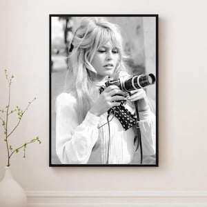 Puede incluir: Fotografía enmarcada en blanco y negro de una mujer sosteniendo una cámara vintage. La mujer tiene el pelo largo y ondulado y lleva una blusa blanca. La cámara tiene una lente larga y una correa de lunares en blanco y negro. La fotografía se muestra en una pared blanca.