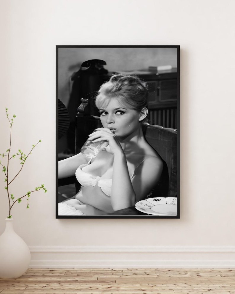 Puede incluir: Impresi&oacute;n enmarcada en blanco y negro de una mujer con sujetador blanco, bebiendo de un vaso. La imagen es una fotograf&iacute;a vintage, con la mujer sentada en una silla. Un plato est&aacute; sobre la mesa. La impresi&oacute;n se muestra en una pared blanca.