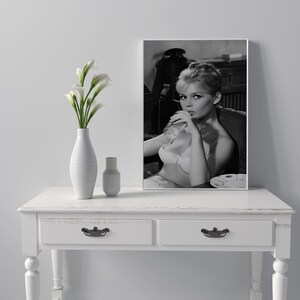 Puede incluir: Una impresi&oacute;n enmarcada en blanco y negro de una mujer en sujetador, sentada en una silla. La impresi&oacute;n se exhibe sobre una mesa blanca, junto a un jarr&oacute;n blanco con flores blancas y una peque&ntilde;a botella blanca.