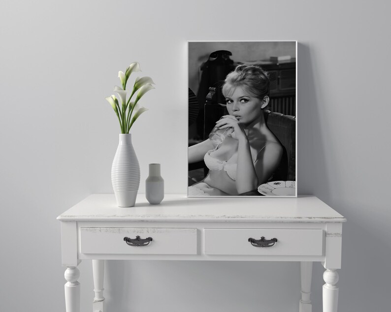 Puede incluir: Una impresi&oacute;n enmarcada en blanco y negro de una mujer en sujetador, sentada en una silla. La impresi&oacute;n se exhibe sobre una mesa blanca, junto a un jarr&oacute;n blanco con flores blancas y una peque&ntilde;a botella blanca.