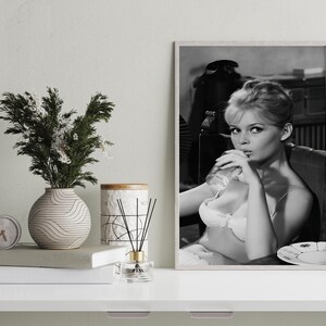 Puede incluir: Impresi&oacute;n en blanco y negro enmarcada de una mujer bebiendo de un vaso. La mujer lleva sujetador y est&aacute; sentada. Un jarr&oacute;n con vegetaci&oacute;n, un reloj, libros y una vela est&aacute;n sobre una superficie blanca.