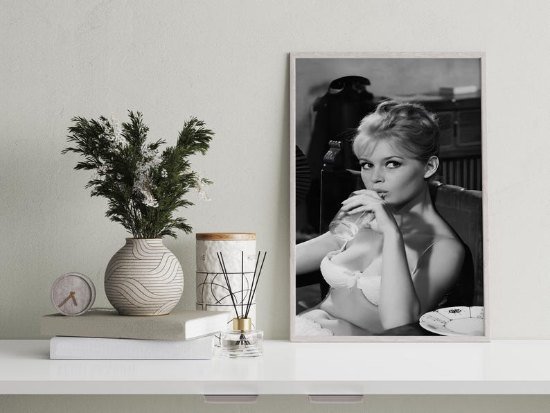 Puede incluir: Impresi&oacute;n en blanco y negro enmarcada de una mujer bebiendo de un vaso. La mujer lleva sujetador y est&aacute; sentada. Un jarr&oacute;n con vegetaci&oacute;n, un reloj, libros y una vela est&aacute;n sobre una superficie blanca.