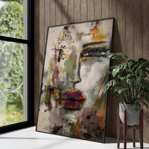 Puede incluir: Impresión artística de un retrato abstracto que representa el rostro de una mujer con labios rojos y ojos expresivos. La obra utiliza una paleta de tonos apagados con toques de color, enmarcada en negro. La pintura se apoya en una pared de madera.