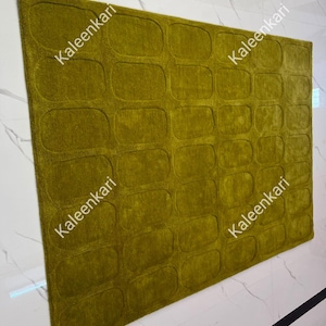 May include: A rectangular area rug in a deep olive green color featuring a repeating pattern of embossed rounded rectangles across its surface. The word Kaleenkari is printed multiple times in white text across the rug's textured fabric.