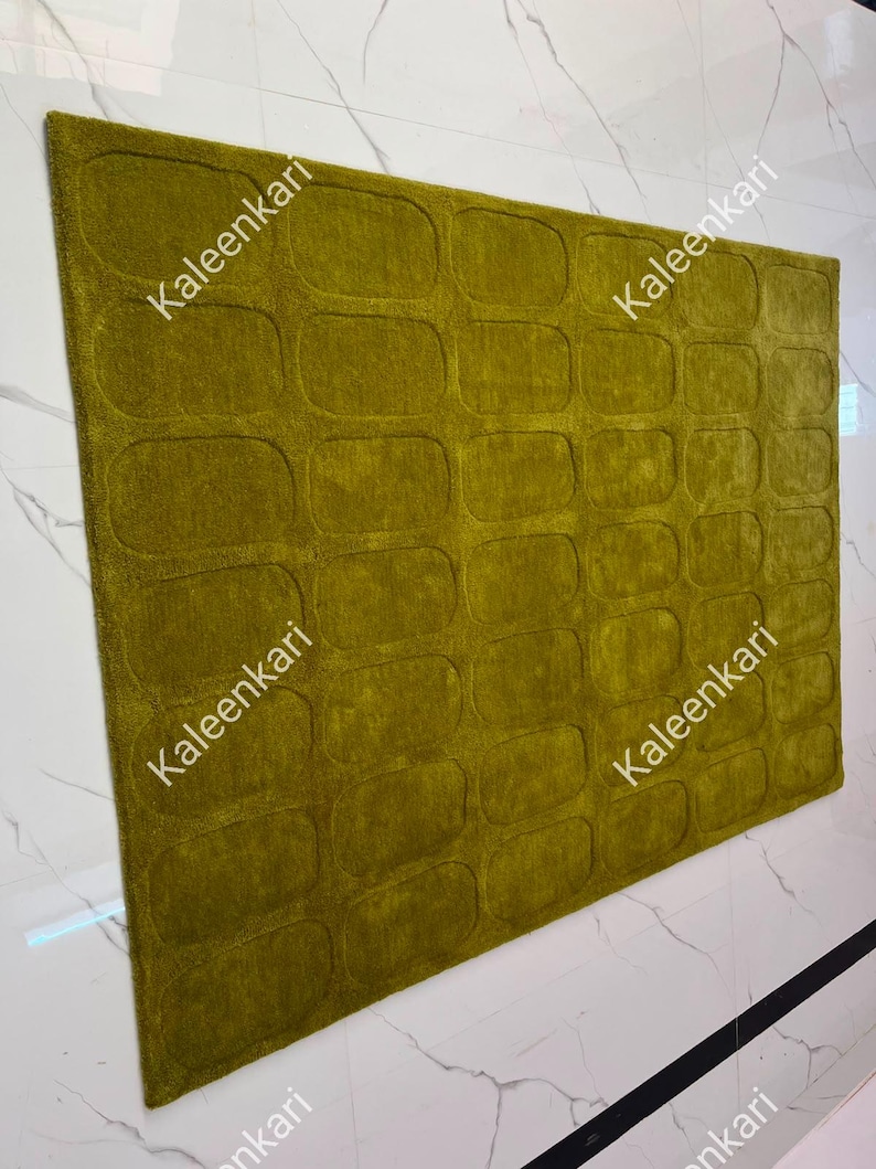 May include: A rectangular area rug in a deep olive green color featuring a repeating pattern of embossed rounded rectangles across its surface. The word Kaleenkari is printed multiple times in white text across the rug's textured fabric.