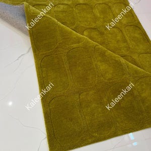 May include: A rectangular olive green rug featuring a repeating pattern of raised, rounded rectangular shapes across its surface. The rug is partially folded back to reveal a light gray backing material. The text Kaleenkari appears on the rug.