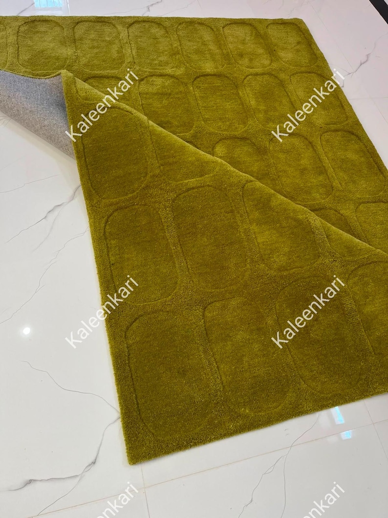 May include: A rectangular olive green rug featuring a repeating pattern of raised, rounded rectangular shapes across its surface. The rug is partially folded back to reveal a light gray backing material. The text Kaleenkari appears on the rug.