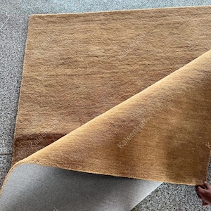 Puede incluir: Una alfombra rectangular marr&oacute;n con una superficie texturizada. La esquina de la alfombra est&aacute; levantada, revelando un reverso gris. La alfombra parece estar hecha de un material suave y tejido, adecuado para una sala de estar o un dormitorio.