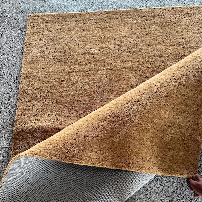 Puede incluir: Una alfombra rectangular marr&oacute;n con una superficie texturizada. La esquina de la alfombra est&aacute; levantada, revelando un reverso gris. La alfombra parece estar hecha de un material suave y tejido, adecuado para una sala de estar o un dormitorio.