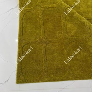 May include: A textured olive green rug featuring a repeating pattern of recessed, rounded rectangular shapes across its surface. The word Kaleenkari is printed in white text multiple times across the rug, indicating the brand name.