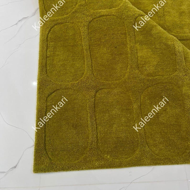 May include: A textured olive green rug featuring a repeating pattern of recessed, rounded rectangular shapes across its surface. The word Kaleenkari is printed in white text multiple times across the rug, indicating the brand name.