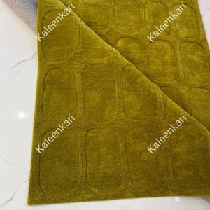 May include: A chartreuse green area rug featuring a repeating pattern of recessed rounded rectangular shapes. The rug is folded over at one corner, revealing a grey backing. The word Kaleenkari is printed multiple times across the surface.