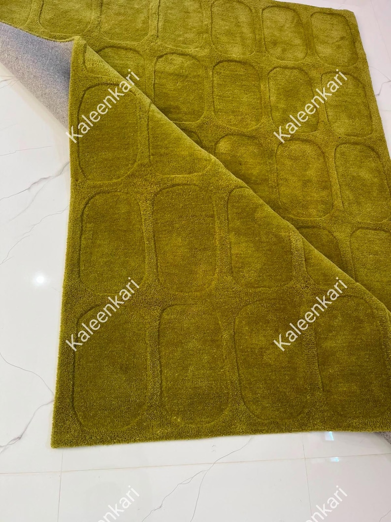 May include: A chartreuse green area rug featuring a repeating pattern of recessed rounded rectangular shapes. The rug is folded over at one corner, revealing a grey backing. The word Kaleenkari is printed multiple times across the surface.
