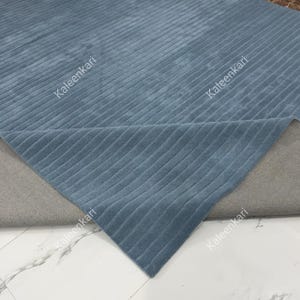 May include: A blue area rug featuring a textured, linear ribbed pattern across its surface. The rug is partially folded, revealing a solid gray backing material. The word Kaleenkari is printed multiple times across the blue surface of the rug.