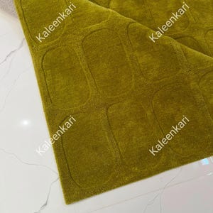 May include: A chartreuse green area rug featuring a repeating pattern of raised, rounded rectangular shapes. The rug is folded at one corner, revealing a light gray backing. The text Kaleenkari is printed multiple times across the surface.