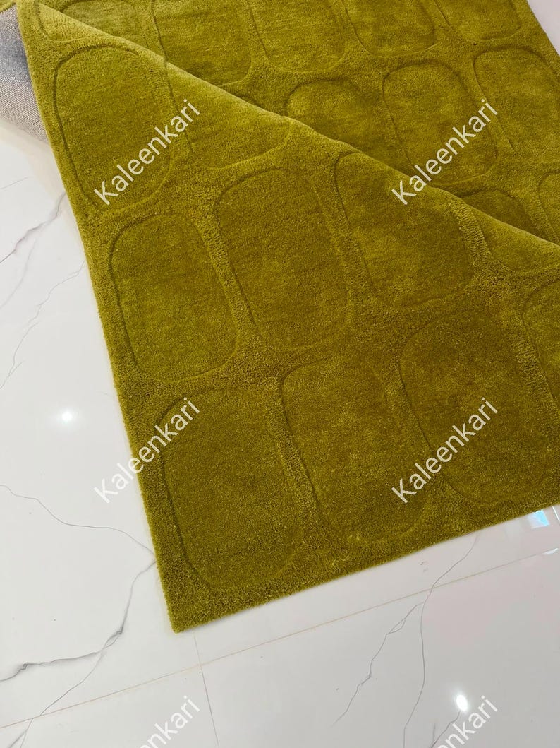 May include: A chartreuse green area rug featuring a repeating pattern of raised, rounded rectangular shapes. The rug is folded at one corner, revealing a light gray backing. The text Kaleenkari is printed multiple times across the surface.