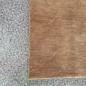 Puede incluir: Primer plano de una alfombra marr&oacute;n con superficie texturizada. La alfombra es rectangular y tiene un aspecto suave y afelpado. La alfombra se coloca junto a una superficie gris moteada.