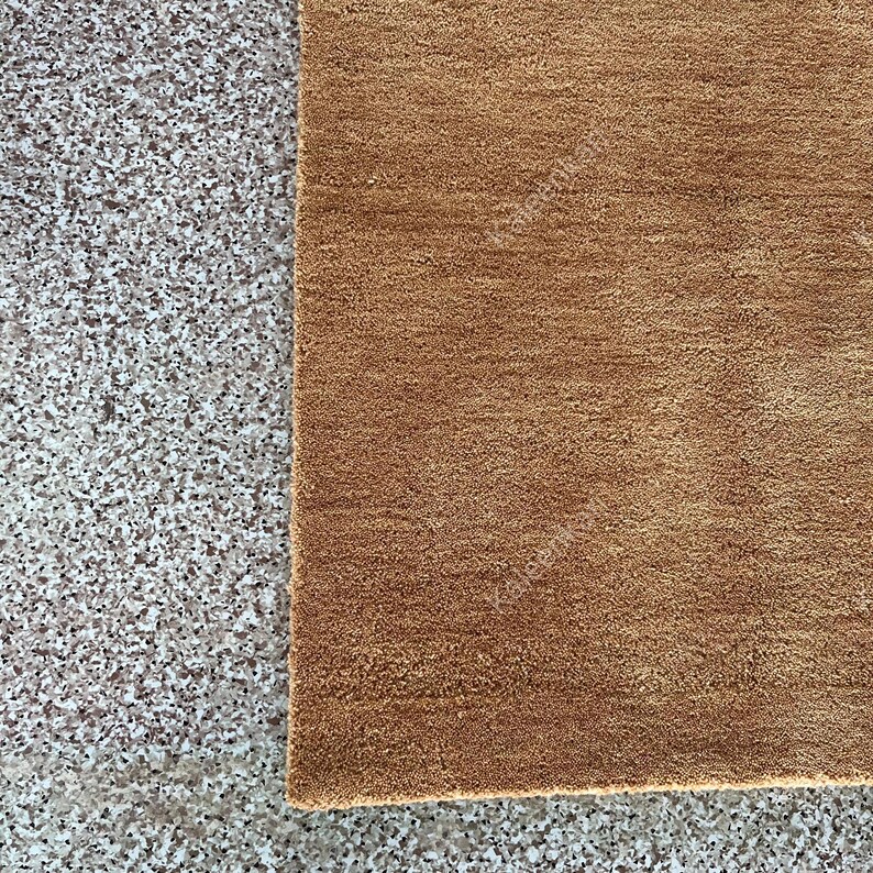 Puede incluir: Primer plano de una alfombra marr&oacute;n con superficie texturizada. La alfombra es rectangular y tiene un aspecto suave y afelpado. La alfombra se coloca junto a una superficie gris moteada.