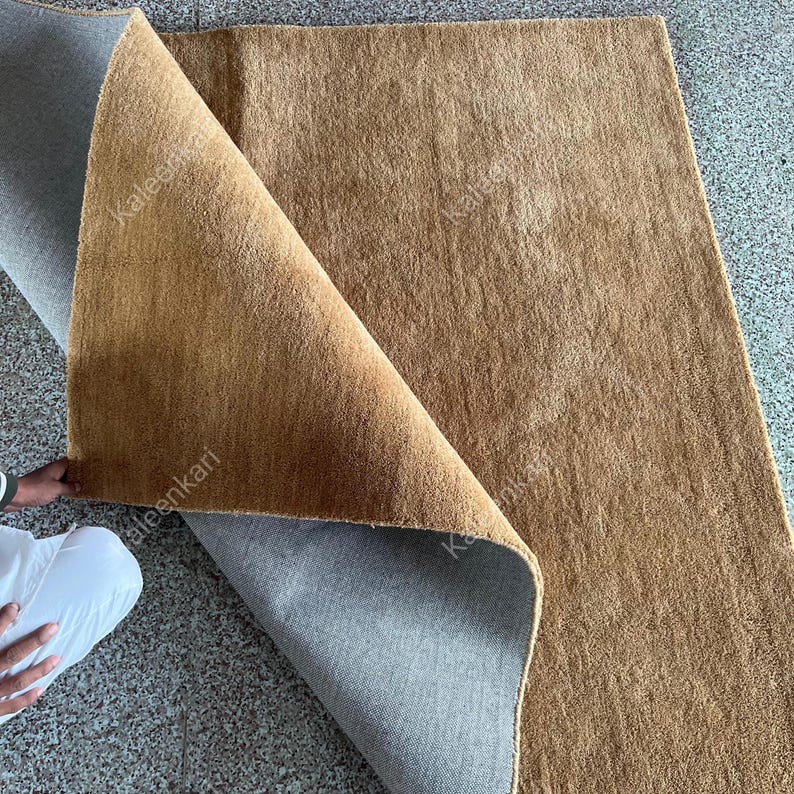 Puede incluir: Una alfombra rectangular marr&oacute;n con una superficie texturizada est&aacute; parcialmente doblada hacia atr&aacute;s, revelando una parte inferior gris. La alfombra parece estar hecha de un material tejido. La alfombra est&aacute; sobre una superficie gris.