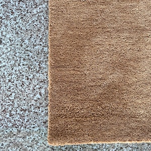 Puede incluir: Primer plano de una alfombra rectangular marr&oacute;n con superficie texturizada. La alfombra est&aacute; hecha de un material suave y tiene una textura ligeramente irregular. La alfombra se coloca junto a una superficie gris moteada.