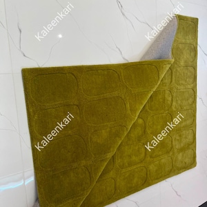 May include: A rectangular olive green rug featuring a repeating pattern of embossed rounded rectangles. The rug is folded over at one corner, revealing a light gray backing material. The surface has a uniform, dense, and soft-textured pile.