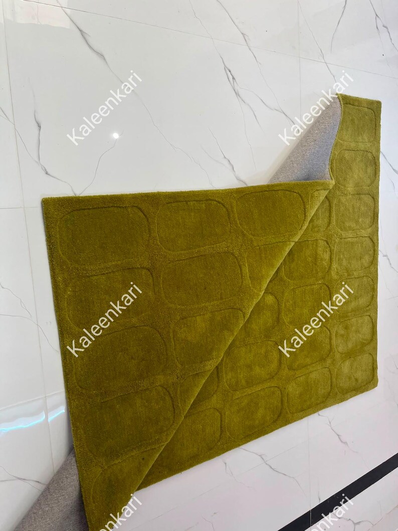 May include: A rectangular olive green rug featuring a repeating pattern of embossed rounded rectangles. The rug is folded over at one corner, revealing a light gray backing material. The surface has a uniform, dense, and soft-textured pile.