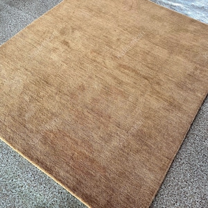 Puede incluir: Una alfombra cuadrada marr&oacute;n con una superficie texturizada. La alfombra es de color liso y parece estar hecha de un material suave. La alfombra est&aacute; colocada sobre una superficie gris moteada.