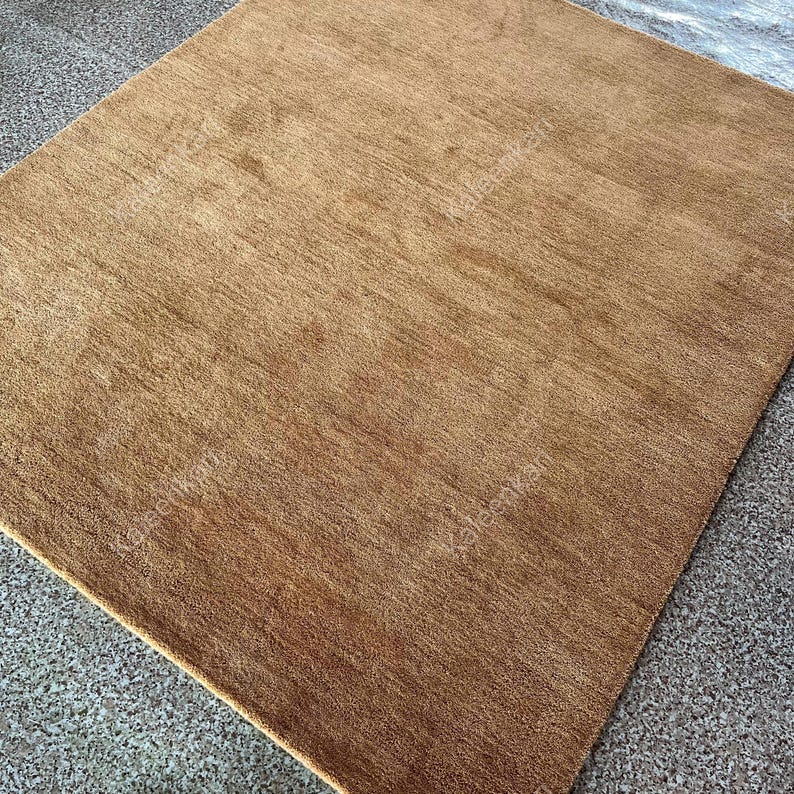 Puede incluir: Una alfombra cuadrada marr&oacute;n con una superficie texturizada. La alfombra es de color liso y parece estar hecha de un material suave. La alfombra est&aacute; colocada sobre una superficie gris moteada.