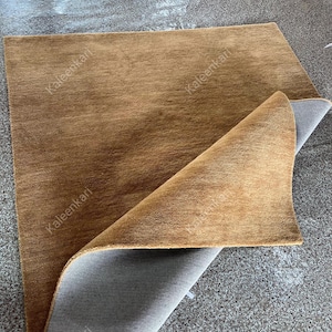 Puede incluir: Una alfombra rectangular marr&oacute;n con una superficie texturizada. La alfombra est&aacute; parcialmente doblada, revelando un reverso gris. El nombre de la marca "Kaleenkari" est&aacute; impreso en la alfombra.