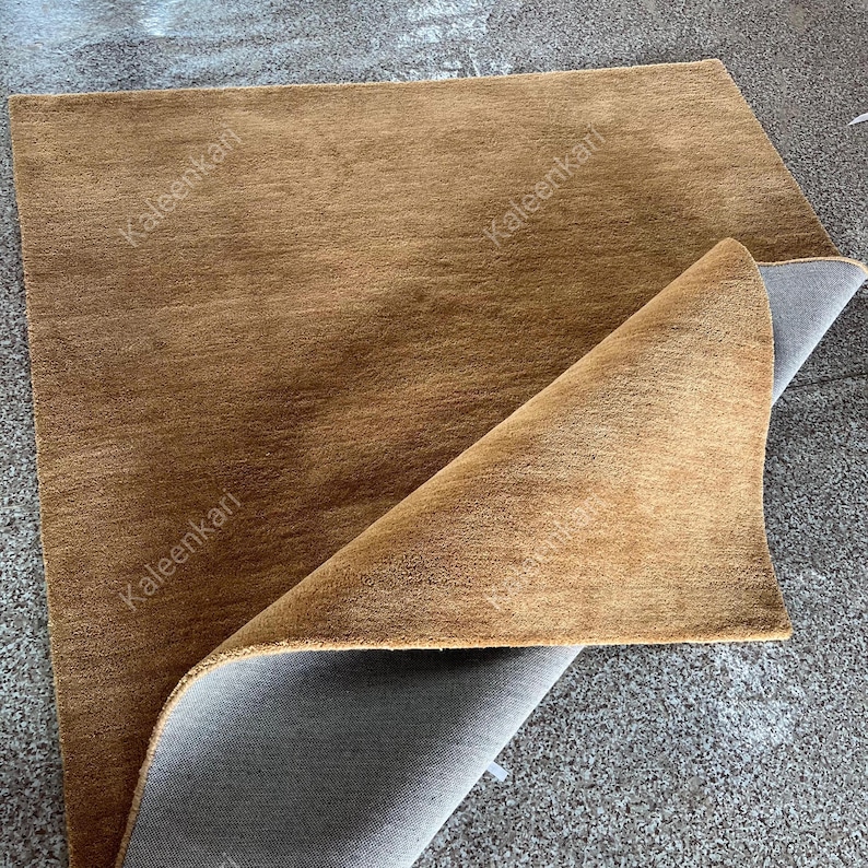 Puede incluir: Una alfombra rectangular marr&oacute;n con una superficie texturizada. La alfombra est&aacute; parcialmente doblada, revelando un reverso gris. El nombre de la marca "Kaleenkari" est&aacute; impreso en la alfombra.