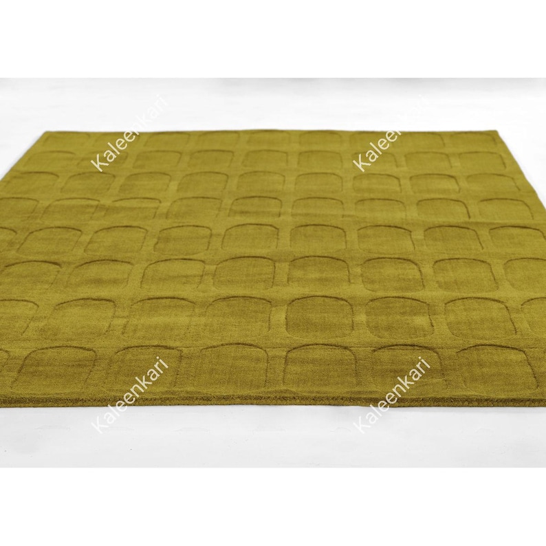May include: A rectangular area rug in a solid olive green color featuring a repeating grid pattern of recessed, rounded square shapes across the entire surface. The word Kaleenkari is printed in white text diagonally across the rug.
