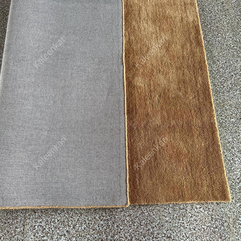 Puede incluir: Primer plano de dos alfombras rectangulares. Una alfombra es de color gris liso y la otra es de color marr&oacute;n liso. Las alfombras est&aacute;n una al lado de la otra, con un borde beige. Las alfombras est&aacute;n sobre una superficie gris.