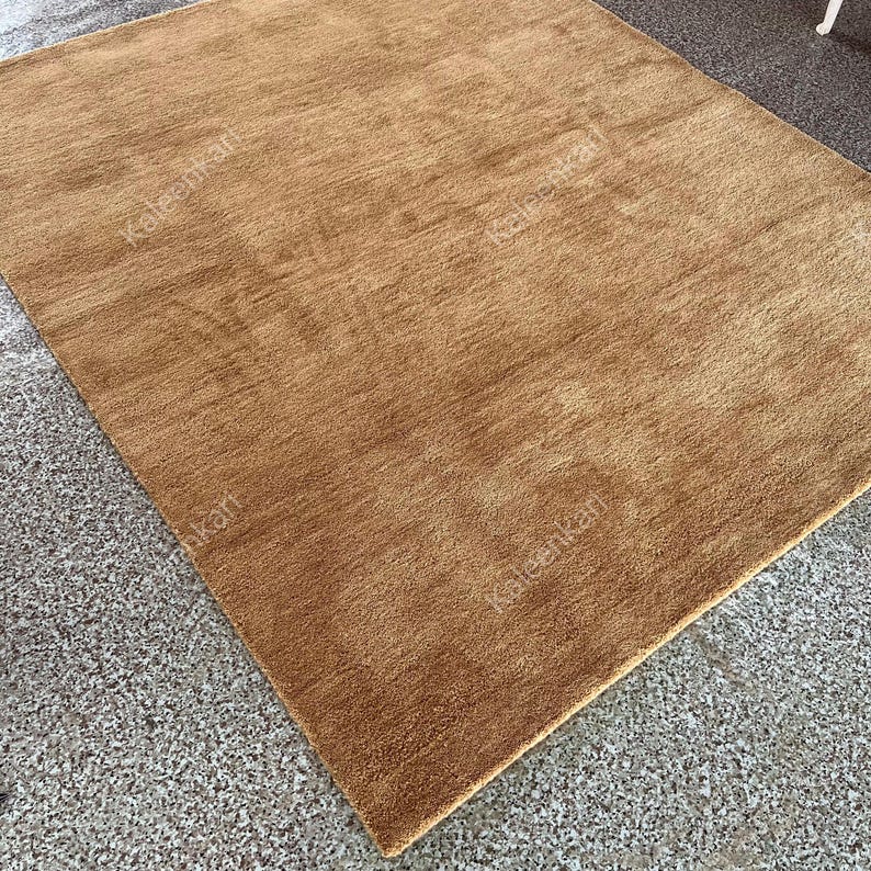 Puede incluir: Una alfombra rectangular de color marr&oacute;n dorado con una superficie texturizada. La alfombra est&aacute; hecha de un material suave y est&aacute; colocada sobre una superficie gris moteada. El color de la alfombra es uniforme.