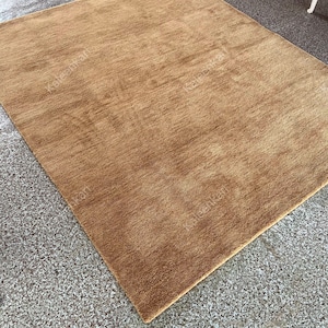 Puede incluir: Una alfombra rectangular de pelo medio en un c&aacute;lido tono marr&oacute;n dorado. La alfombra tiene un aspecto suave y texturizado, con sutiles variaciones de color. La alfombra est&aacute; colocada sobre una superficie gris moteada.