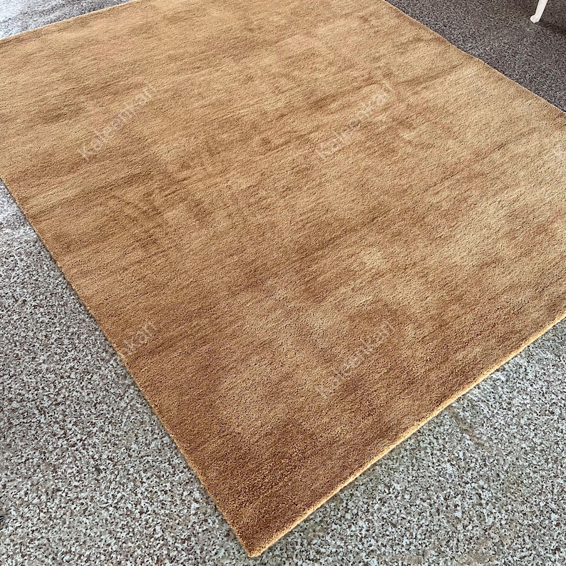 Puede incluir: Una alfombra rectangular de pelo medio en un c&aacute;lido tono marr&oacute;n dorado. La alfombra tiene un aspecto suave y texturizado, con sutiles variaciones de color. La alfombra est&aacute; colocada sobre una superficie gris moteada.