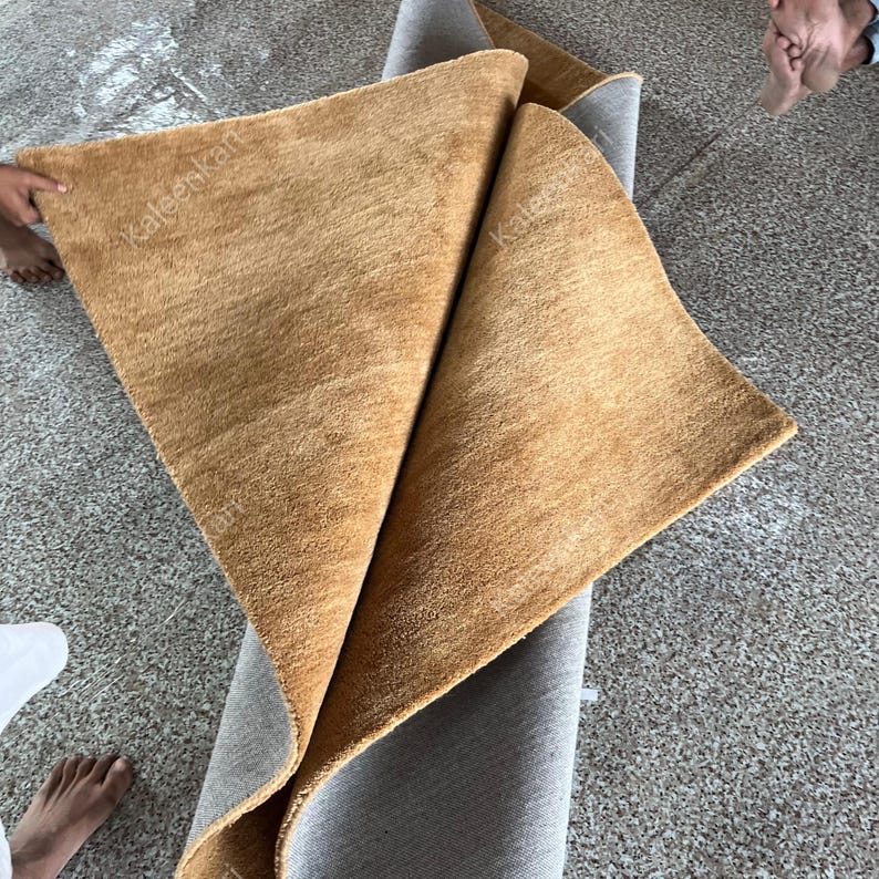 Puede incluir: Una alfombra rectangular doblada con una superficie texturizada de color marr&oacute;n dorado. La alfombra est&aacute; parcialmente enrollada, revelando un respaldo gris. La imagen muestra la textura suave y afelpada de la alfombra y las variaciones de color.