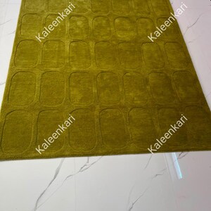 May include: A rectangular olive green rug featuring a repeating pattern of raised, rounded rectangular shapes across its surface. The word Kaleenkari is printed multiple times in white text across the rug, which has a textured, plush pile.