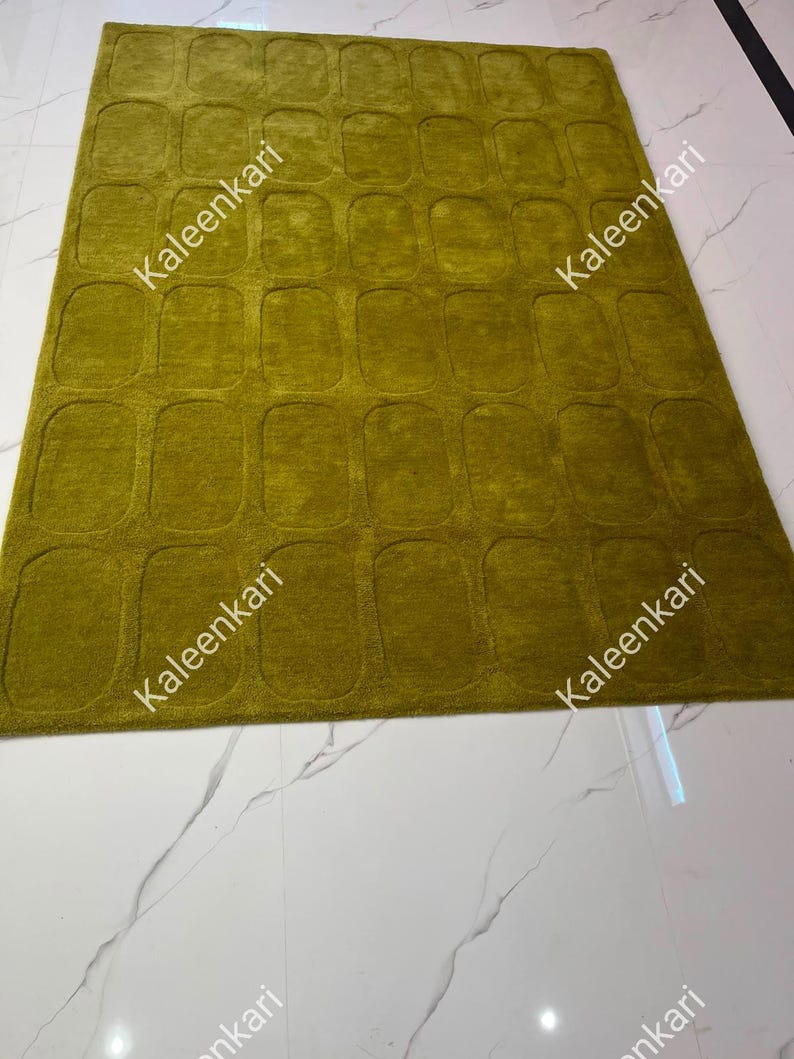 May include: A rectangular olive green rug featuring a repeating pattern of raised, rounded rectangular shapes across its surface. The word Kaleenkari is printed multiple times in white text across the rug, which has a textured, plush pile.