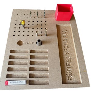 May include: A wooden tool organizer with various drill bits, collets, and a red container. The text "Thickness Gauge" and "Collets" are visible. A label reads "30 Deg V-Bit 3 Flute".