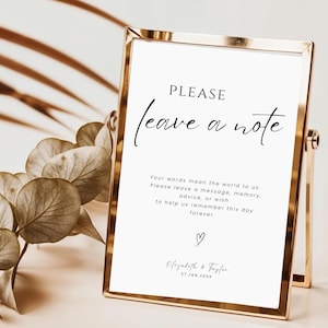 May include: A gold-toned metal frame holding a white sign with black text that reads: PLEASE leave a note. Your words mean the world to us. Please leave a message, memory, advice, or wish to help us remember this day forever. Elizabeth & Taylor 27.JAN.20XX.