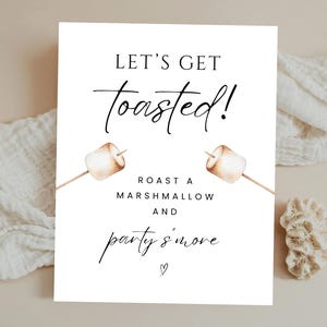Peut inclure: Une affiche rectangulaire blanche avec le texte "LET'S GET toasted!" en écriture noire. En dessous, on peut lire "ROAST A MARSHMALLOW AND party s'more" avec des illustrations à l'aquarelle de guimauves grillées sur des bâtons.
