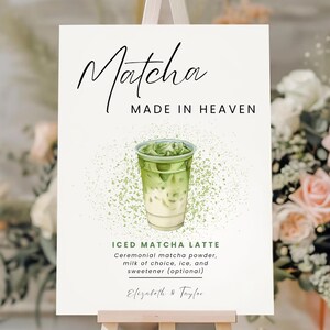Puede incluir: Un letrero rectangular blanco sobre un caballete de madera con el texto Matcha MADE IN HEAVEN, una ilustración de un latte de matcha helado, el texto ICED MATCHA LATTE, una lista de ingredientes y los nombres Elizabeth & Taylor en negro.