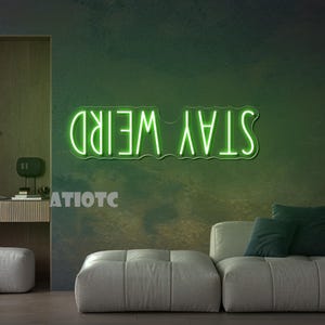 May include: A neon green sign that reads "STAY WEIRD" in a modern font. The sign is mounted on a textured, dark green wall. The image also shows a modern living room with a gray sofa and green pillows.